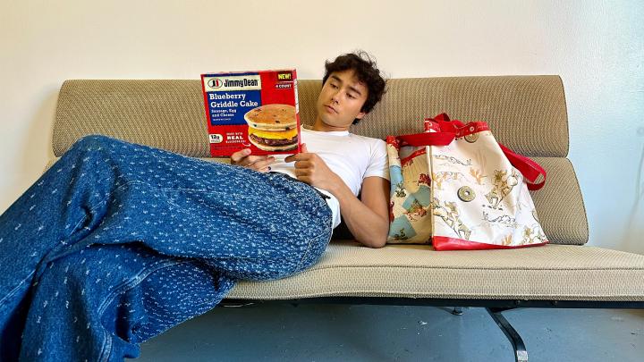 Person sitting on couch with blue blanket holding a box of Jimmy Dean, next to them on couch is Jimmy Dean Griddle Bag